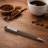 A brown pen with a metallic tip positioned on a wooden surface, surrounded by coffee grounds, beans, and a cup of coffee.