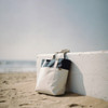 A beach tote bag in cream and navy rests against a white wall on sandy shore, with the ocean in the background.