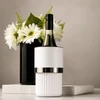 A white wine bottle cooler with a silver band, placed on a marble surface beside a black vase with white flowers.