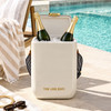 A white cooler bag holds two bottles of champagne surrounded by ice, set beside a pool with a tan-striped towel.