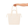 A cream tote bag with two handles and a front pocket, held by a hand with red nails. It has a logo.