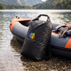 A black 20L dry bag with a logo, resting beside an inflatable kayak on a gravel shore near water.