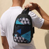 A crossbody bag featuring a geometric pattern in black, turquoise, and blue, held by a person. It has a logo.