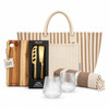 A striped tote bag, a wooden cheese board, a cheese knife set, two clear glasses, and a patterned blanket.