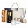 A striped tote bag contains a cheese set, a wooden cutting board, two glass tumblers, and a striped blanket.
