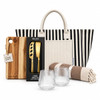 A striped tote bag with a wooden cheese board set, two glass tumblers, and a patterned blanket.