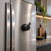 A compact black speaker magnetically attached to a stainless steel fridge in a modern kitchen setting.