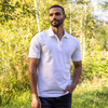 A men's polo shirt in white, featuring a collar and short sleeves, displayed in a natural outdoor setting.