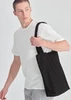 A man in a white T-shirt carries a black canvas tote bag with a shoulder strap. The bag has a simple design.