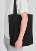 A black canvas tote bag is held by a person wearing a white shirt and grey pants. The bag has a simple design.