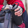 A drink bottle in matte black, featuring a lid with a mouthpiece and a handle, held by a gloved hand next to a backpack.