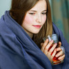 A young woman wrapped in a navy fleece blanket holds a glass mug, sitting in a cozy indoor setting.