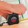 A black tape measure with a logo, being used to measure wood, with a pencil in the other hand.