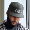A dark green cap with a logo on the front, worn by a person with a beard, standing indoors.