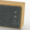 A bamboo Bluetooth speaker with a grey fabric front, featuring control buttons and a logo.