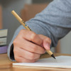 A wooden pen with a black tip is held over a page, with books stacked in the background. It has a logo on its side.