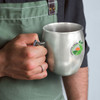 A stainless steel mug with a handle, featuring a logo, held by a person in an apron.