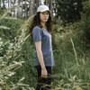 A young woman wearing a grey TRENDSWEAR Carmen women's T-shirt and a white cap stands in a grassy field.