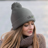 A grey knitted beanie featuring a pom-pom on top and a logo, worn by a woman with long hair.