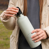 A drink bottle in a soft green colour with a carry lid, held by a person wearing a light jacket in an outdoor setting.