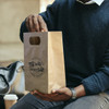 A medium die cut paper bag in brown, featuring a logo, held by a person with a black sweater.