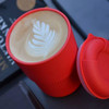 A reusable coffee cup in bright red with a latte featuring a leaf pattern, accompanied by a matching lid.