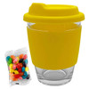 A clear glass coffee cup with a yellow silicone lid and sleeve, accompanied by a bag of colourful jelly beans.