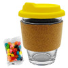 A glass coffee cup with a cork band and a yellow lid, accompanied by a pack of colourful jelly beans.