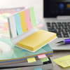 A mini sticky note pad with yellow notes and assorted pastel-coloured flags, set on a desk with a laptop and books.
