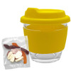 A clear glass coffee cup with a yellow silicone lid and sleeve, accompanied by a bag of mixed lollies.