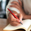 A red metal pen held by a hand over an open notebook, featuring a branding logo on the pen.