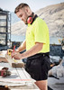 A man wearing a high-visibility yellow polo shirt and black rugby shorts measures wood on a construction site.
