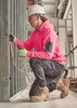 A person in a bright pink water-resistant hoodie and grey work pants kneeling, working on electrical wires in a construction setting.