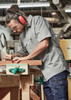 A lightweight grey short sleeve work shirt worn by a man using a pencil on wood in a workshop. The shirt has a logo.