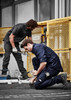 Two men in dark work shirts and pants engage in plumbing tasks outdoors near a yellow fence.