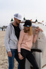 A grey hoodie and a black snapback cap on a male model, alongside a female model in a pink hoodie and black cap, both at the beach.
