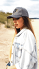 A grey cap worn by a woman with long hair, standing on a sandy beach, wearing a denim jacket.