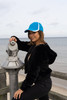 A woman wears a blue cap with white inserts on a seaside pier, posing beside a viewing telescope.