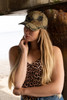 A cap featuring leaf print camouflage design, worn by a model with long hair, set against a natural backdrop.