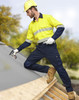 A man wearing a yellow and navy utility shirt and navy cargo pants, standing on a roof, holding a tool.