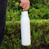 A drink bottle in white with a metal loop, featuring a sleek design, held by a person against a green background.