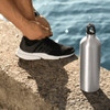 A silver aluminium sports bottle with a clip top beside a person lacing their shoes on a stone surface.