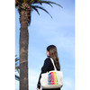 A person holding a multi-coloured zippered cotton canvas tote bag stands near a palm tree against a blue sky.