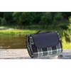 A picnic rug featuring a black and white plaid pattern, neatly rolled with a carrying handle, beside a river.
