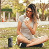 A waterproof Bluetooth speaker in black sits on the grass beside a woman using her smartphone outdoors.