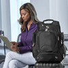 A black checkpoint-friendly backpack with multiple compartments sits beside a woman using a tablet.