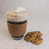 A reusable coffee cup with a cork band and a light-colored lid sits beside a few chocolate chip cookies.