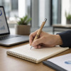 A hand holds a tan Napier Paper Pen over a notepad on a wooden desk, with a laptop and plants in the background.