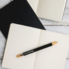 A black aluminium pen rests on an open cream notebook, with a black notepad also visible in the background.