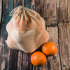 A mesh produce bag in light beige color with two oranges beside it on a wooden surface.